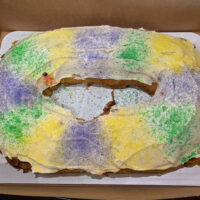 Large, 9 by 13 oval shaped king cake with buttercream frosting and yellow, green, and purple sanding sugar, and a small plastic king cake baby.